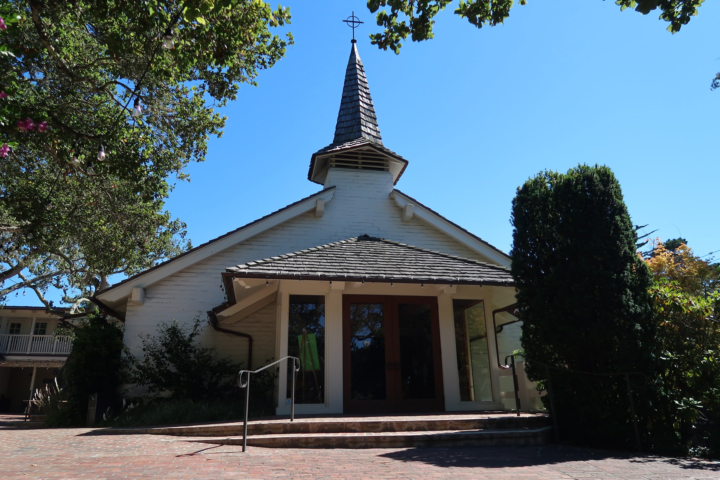View of a small white chapel with a pointed steeple and a cross on top, surrounded by green trees under a clear blue sky.