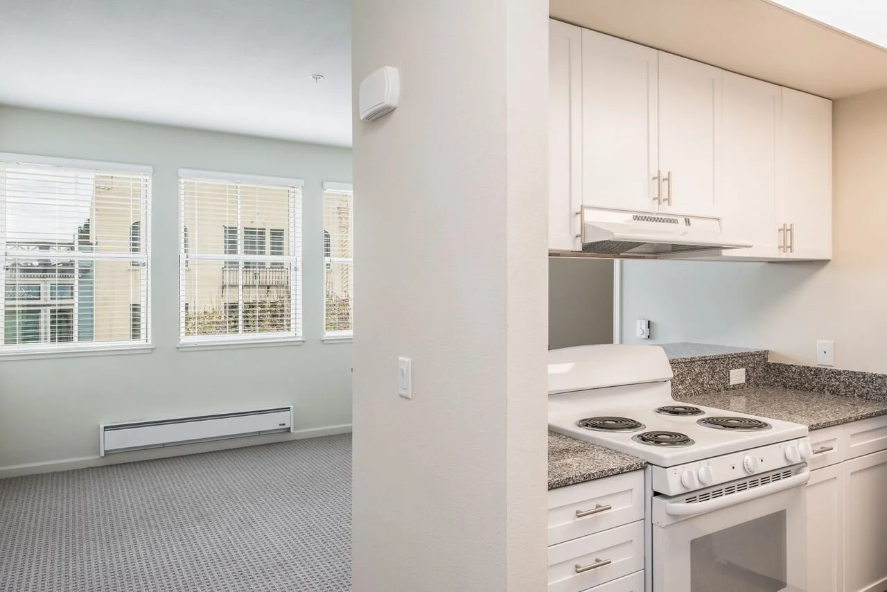 Interior view of a modern apartment with a bright living room featuring three large windows with white blinds and a baseboard heater underneath. The kitchen is partially visible with white cabinets, granite countertops, and a white electric stove wit