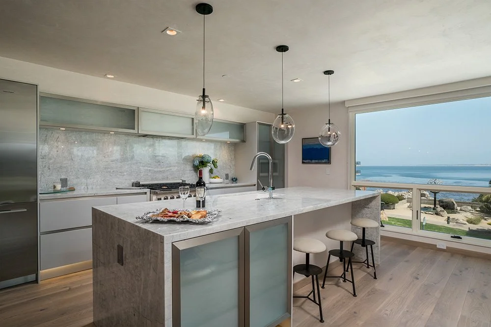 Modern kitchen with a marble island, bar stools, pendant lighting, and a view of the beach and ocean through large windows.