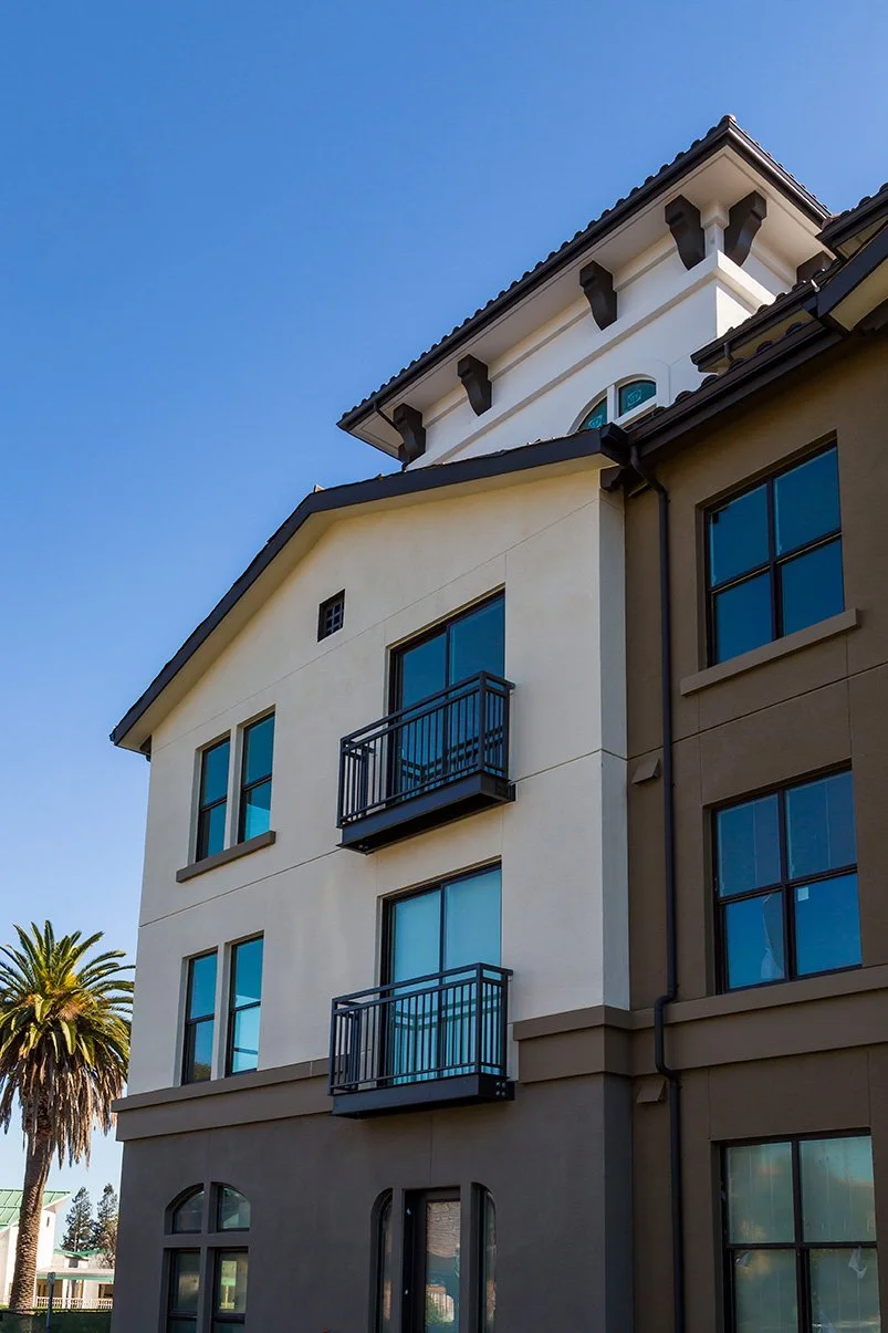 A multi-story modern residential building with beige and white exterior walls, black window frames, small balconies, and a tall palm tree nearby, under a clear blue sky.