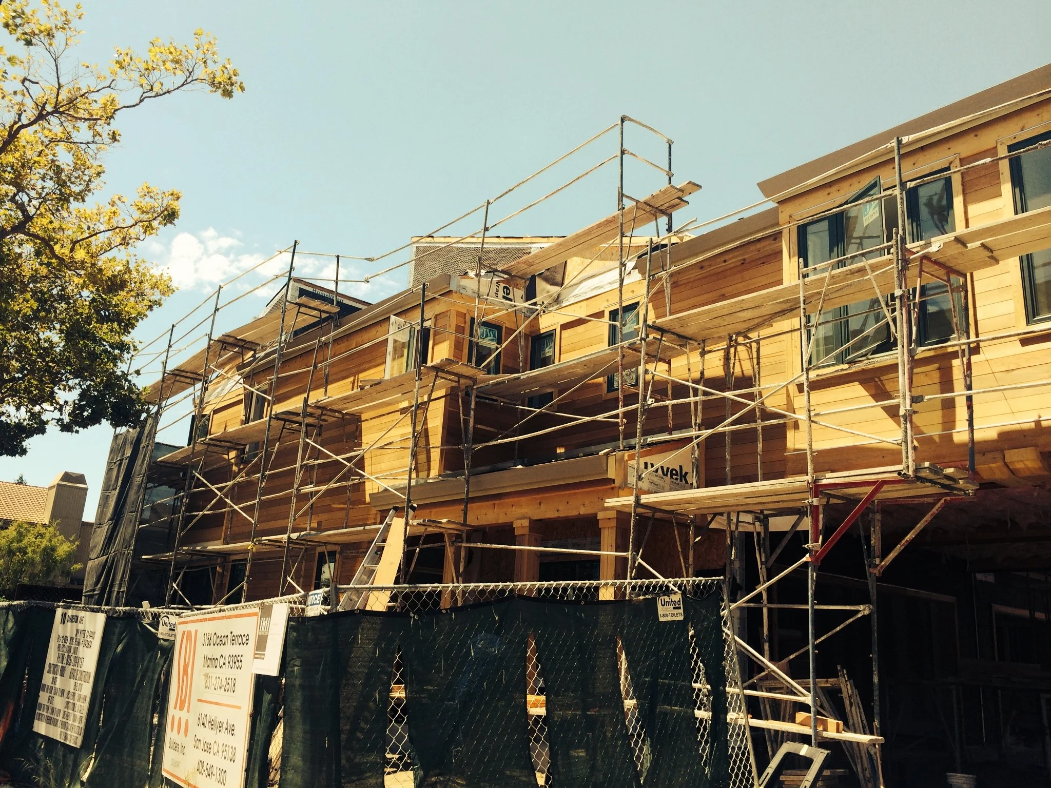 Wooden multi-story building under construction with scaffolding surrounding it, construction signs and a chain-link fence in the foreground, a tree on the left, and a clear blue sky above.