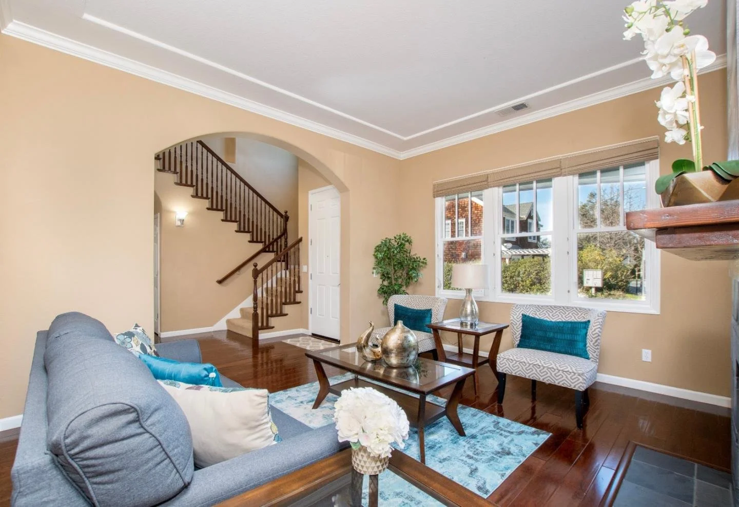 Living room with beige walls, hardwood floors, a blue sofa with pillows, a wooden coffee table with decorative vases, two patterned armchairs with teal pillows, a large window with beige blinds, a tall potted plant, a table lamp, and a wall-mounted s