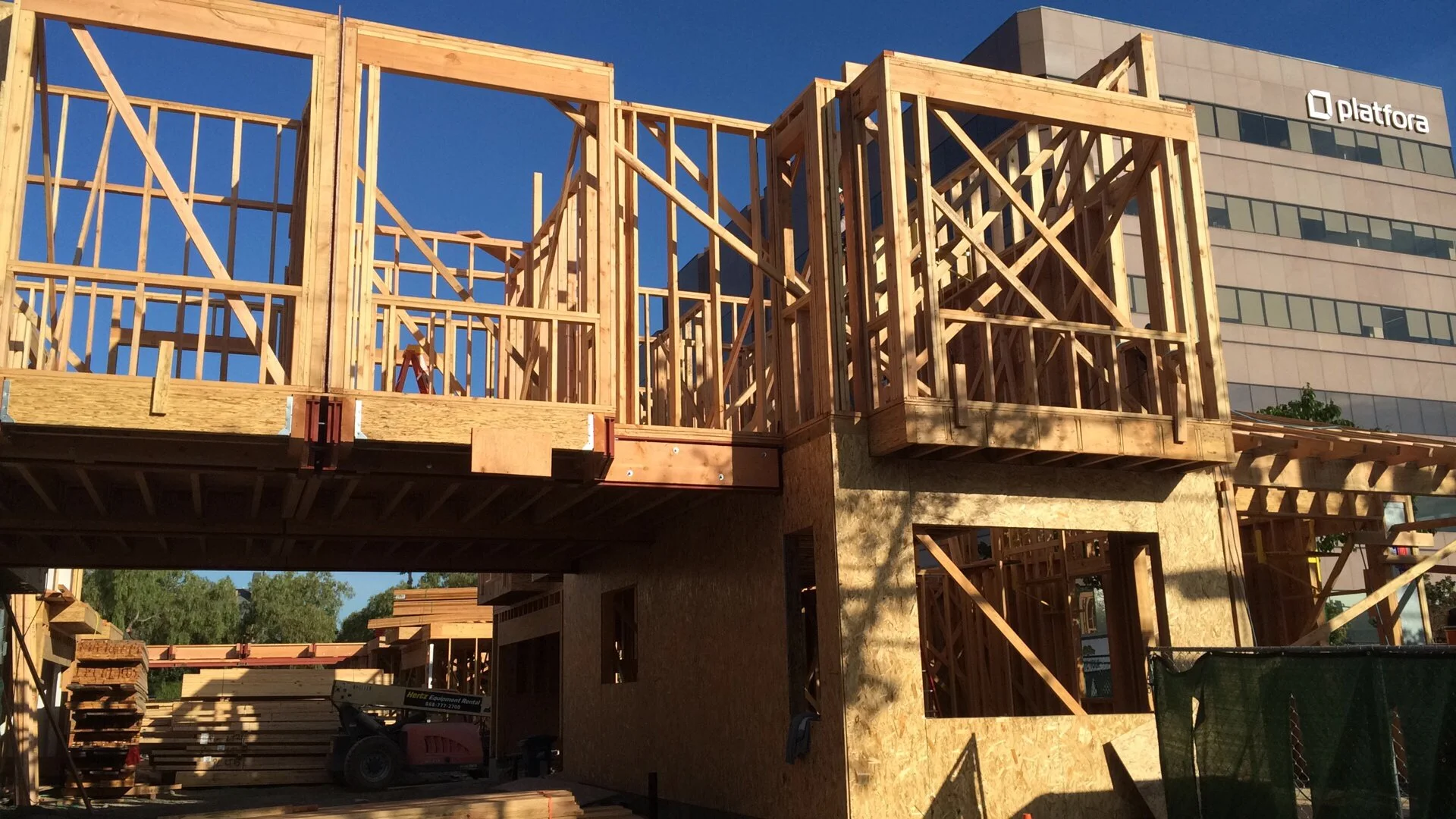 Wooden framing for a building under construction with a second story, set against a blue sky, with a modern building and trees in the background.