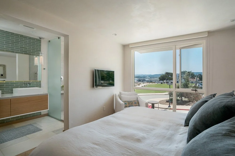 Hotel room with a bed, a TV, a sitting chair, and a large window showing an outdoor view of a parking lot, trees, and distant hills.