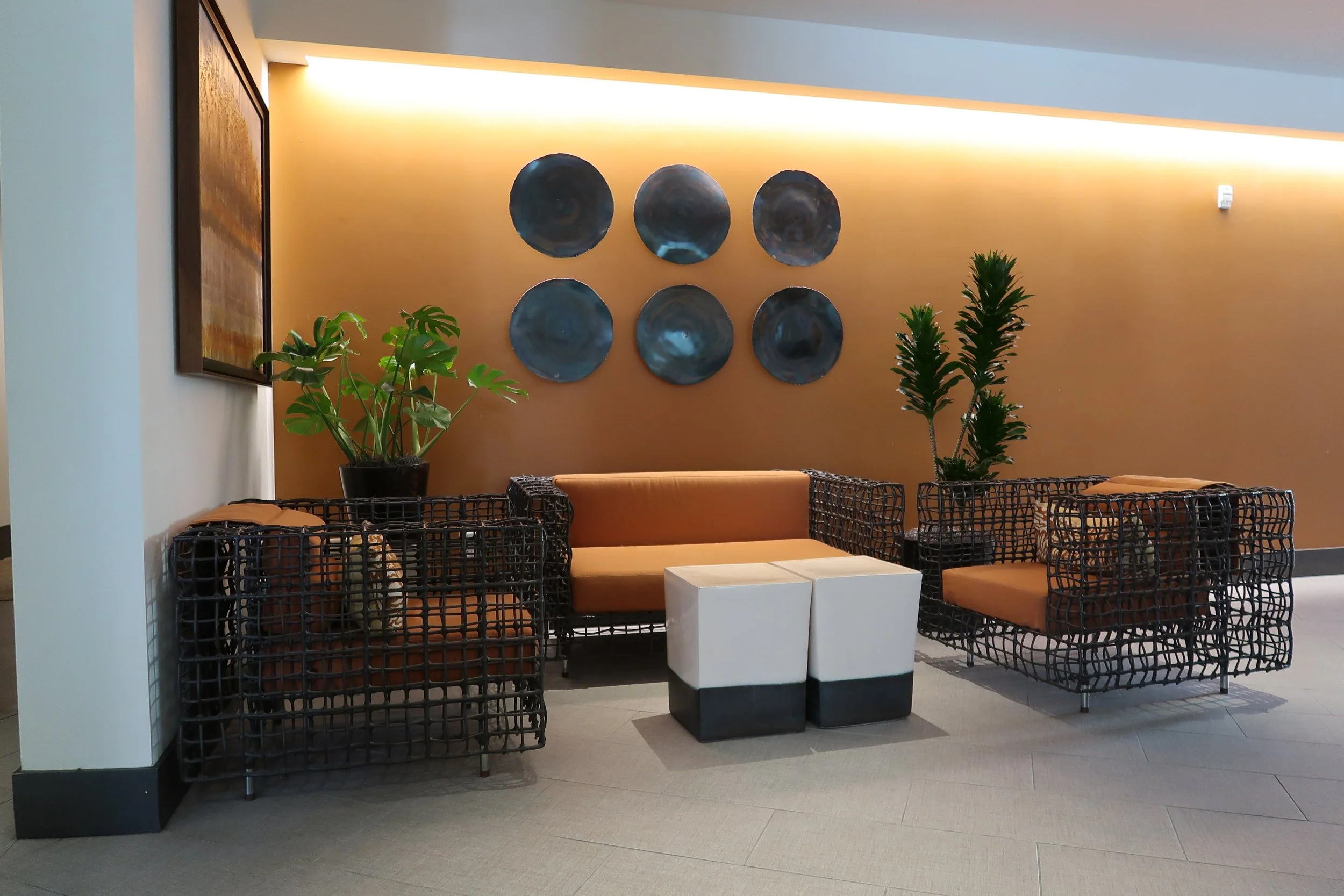 Modern hotel lobby with orange cushioned seating, black woven wicker chairs, small white side tables, potted plants, and decorative wall art on an orange accent wall.