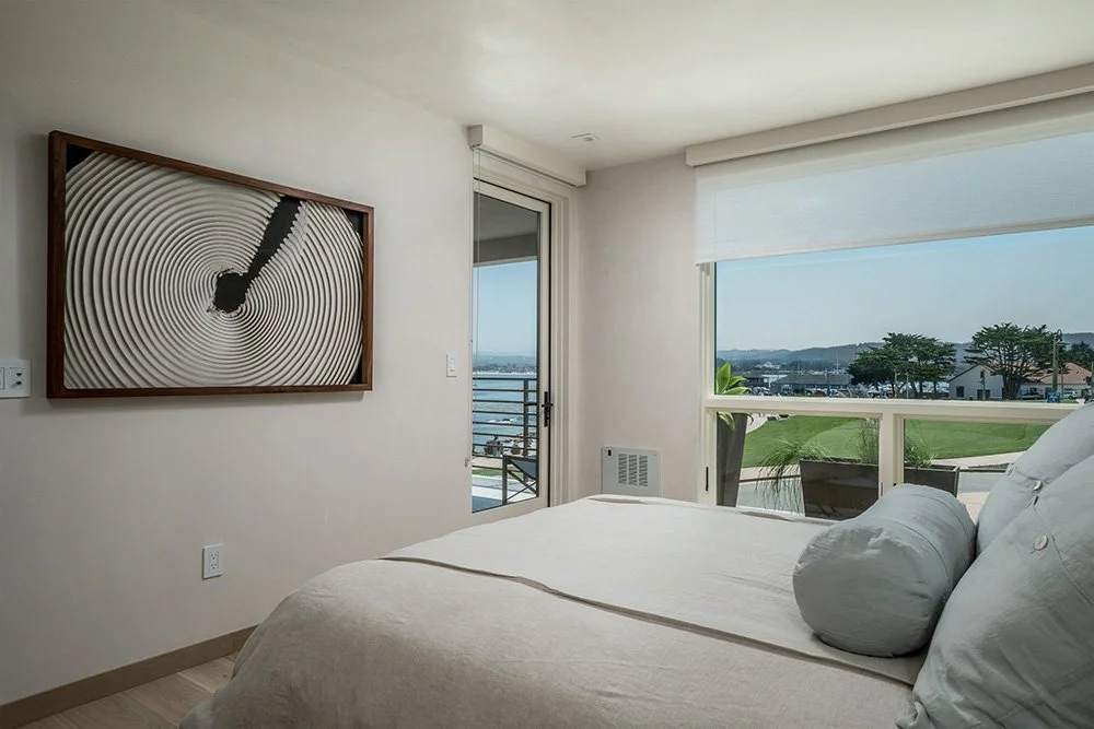 Bedroom with white bedding, large window with view of green lawns and distant buildings, and abstract wall art.