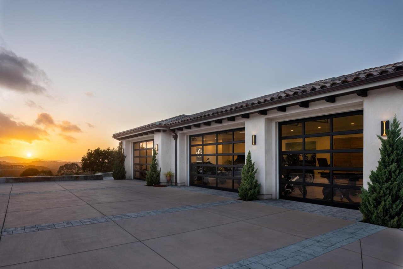 A modern house garage with three glass-paneled rolling doors, some greenery, and a view of the sunset in the background.