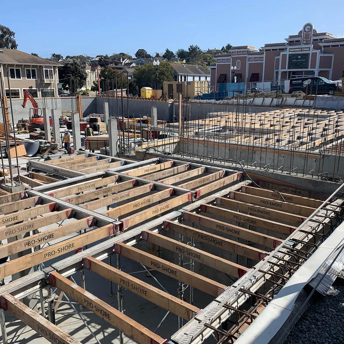 Construction site with wooden frames labeled 'Pro-Shore' being built for a new structure, workers in safety gear, city buildings in the background, clear blue sky.