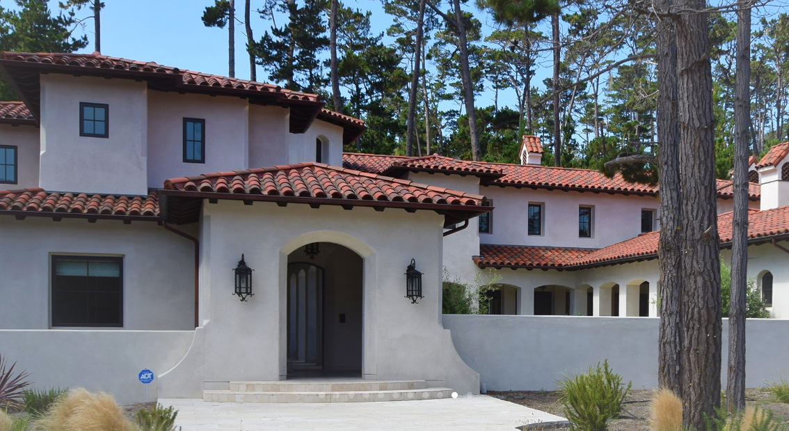 A house with white stucco walls and a red tile roof, surrounded by trees.