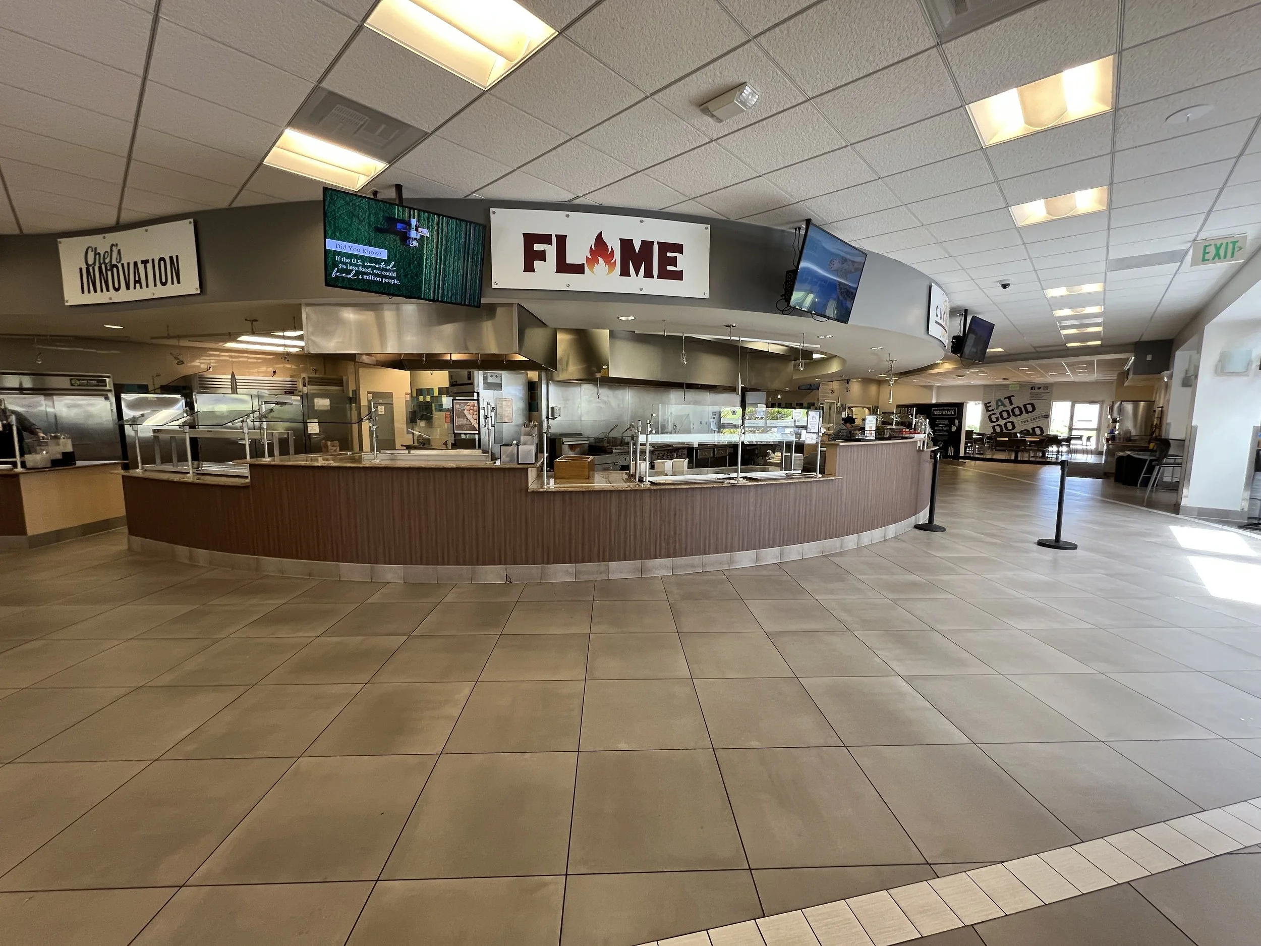 Interior of a restaurant with a curved counter, TV screens, and signs including "Chef's Innovation" and "Flame," with empty tables and chairs.