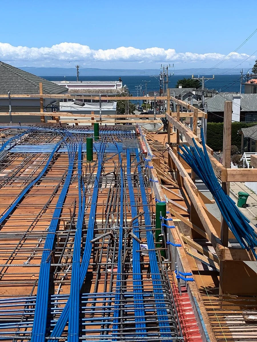 Construction site on a building with exposed blue electrical conduits, wooden framing, and a view of a water body and distant land in the background.