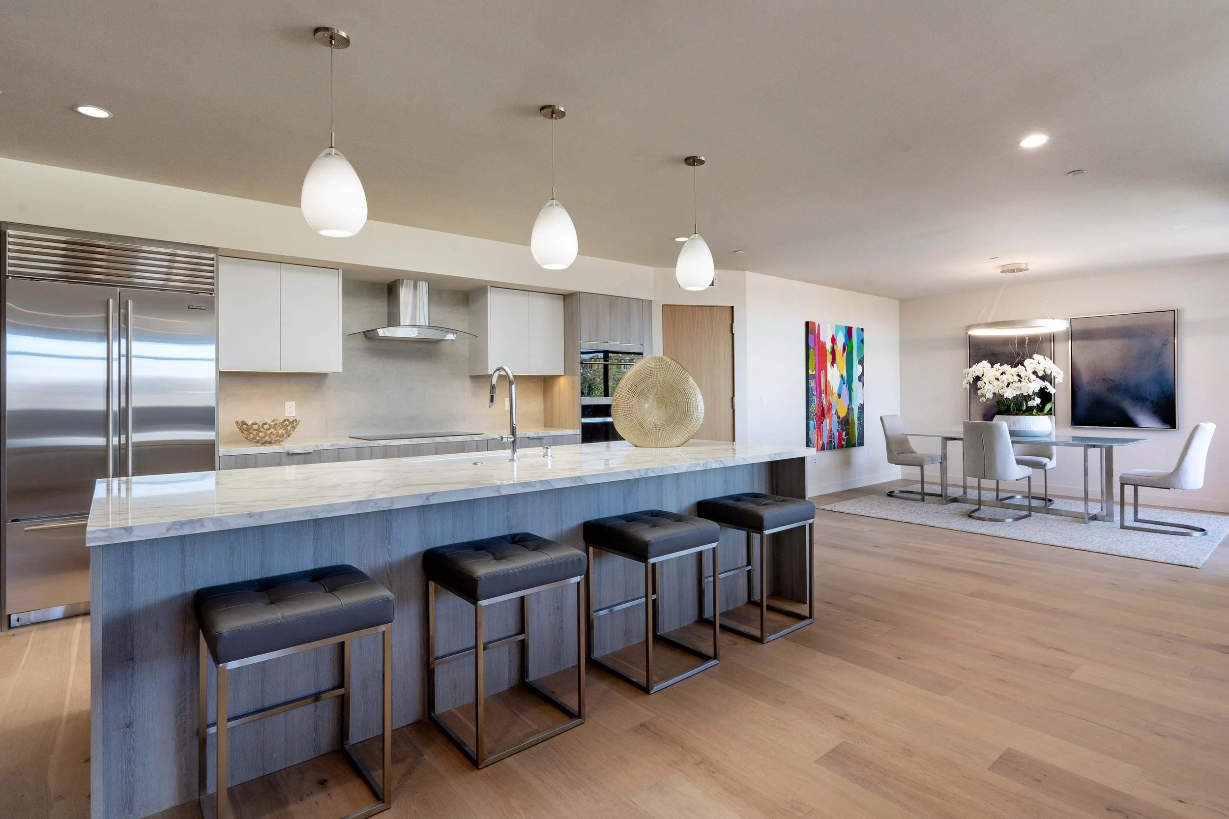 Modern kitchen and dining area with pendant lights, a marble-topped island with four black barstools, and a dining table with four chairs. Artwork and white flowers decorate the space.