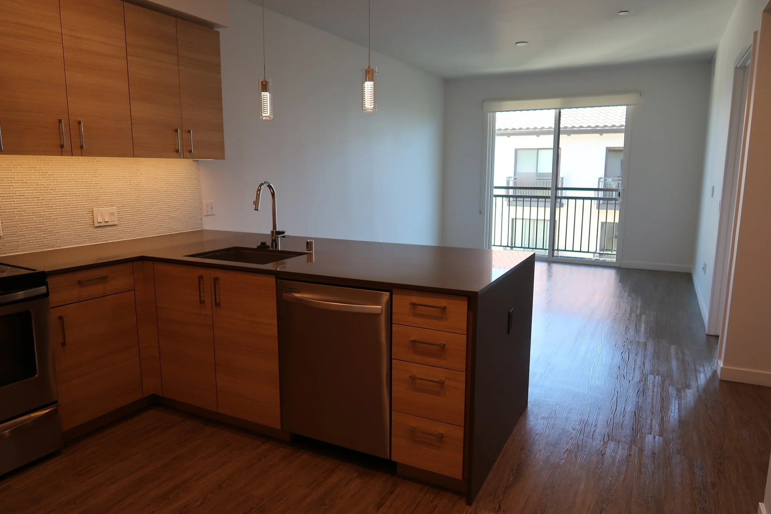 Open-plan kitchen and living room area with wooden cabinets, a kitchen island, hardwood floors, and a sliding glass door leading to a balcony.