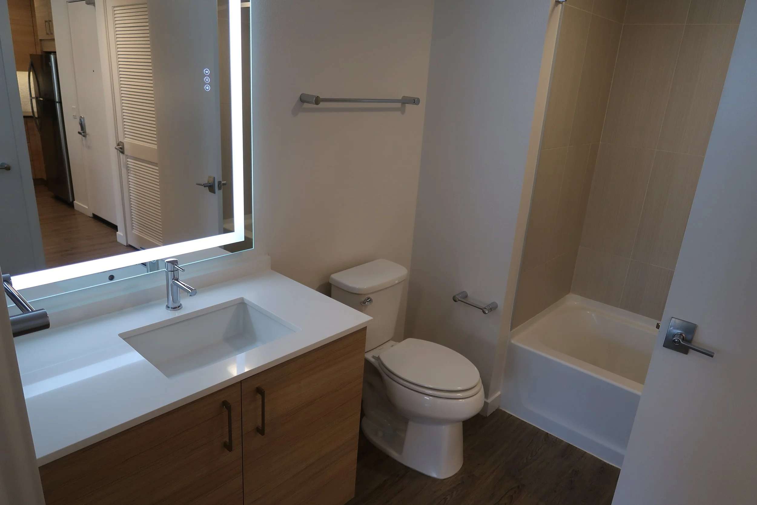 A modern bathroom with a white sink and wooden vanity, a toilet, a bathtub with beige tiles, a large illuminated mirror, and a view into a kitchen area with a refrigerator.