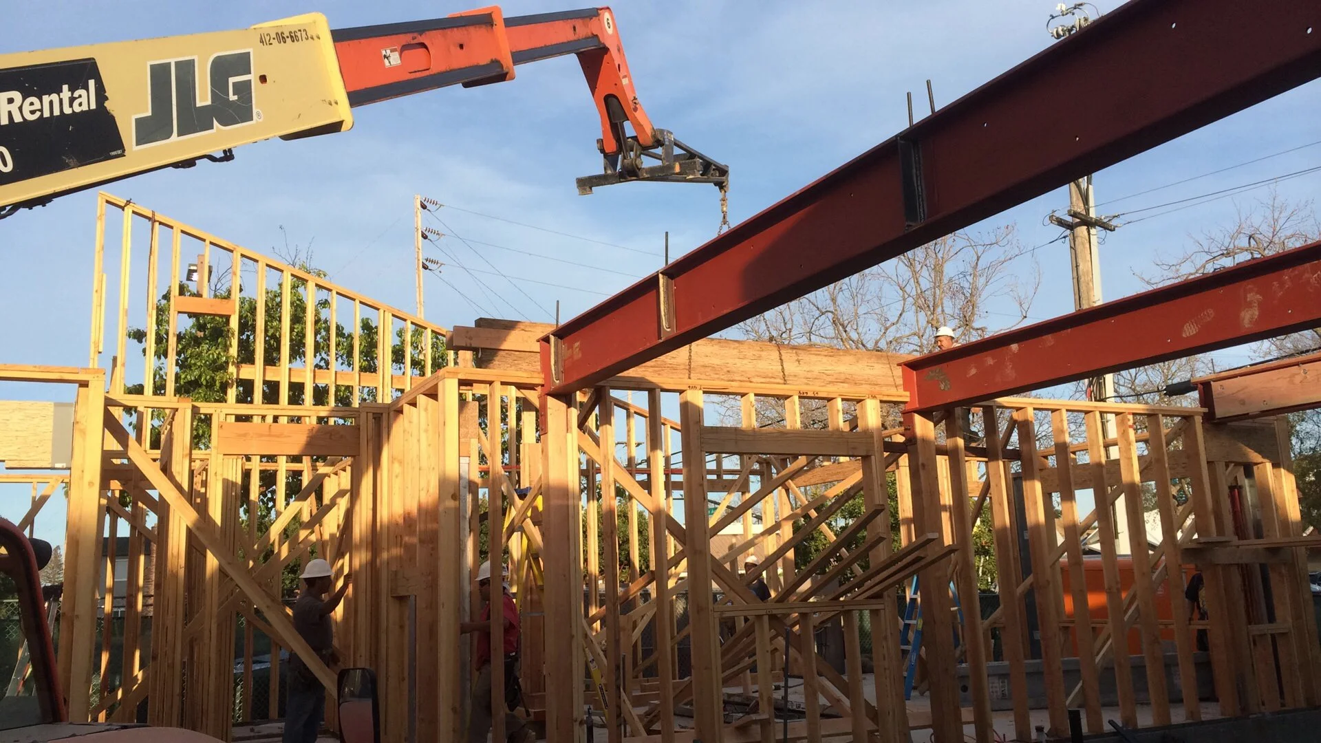 Construction site with wooden framing for a building, a crane lifting steel beams, and workers wearing helmets working on the structure under a clear sky.