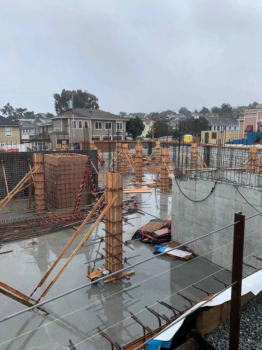 Construction site with concrete foundation, wooden support beams, and rebar, wet and rainy, in a residential neighborhood.