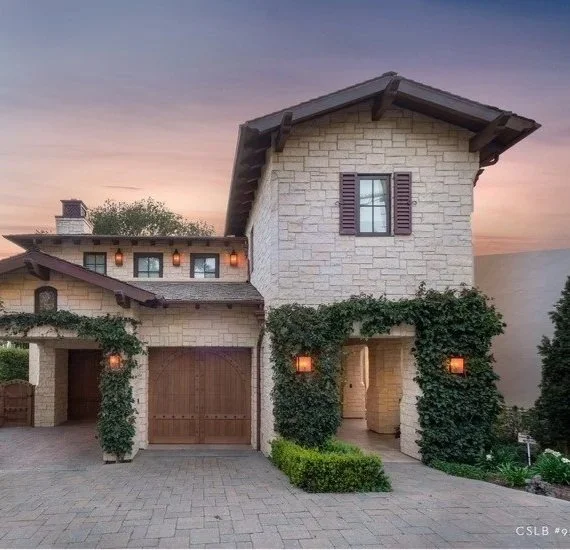 Front of a stone house with a two-car garage, climbing plants, a wooden door, and a small window with shutters at sunset.