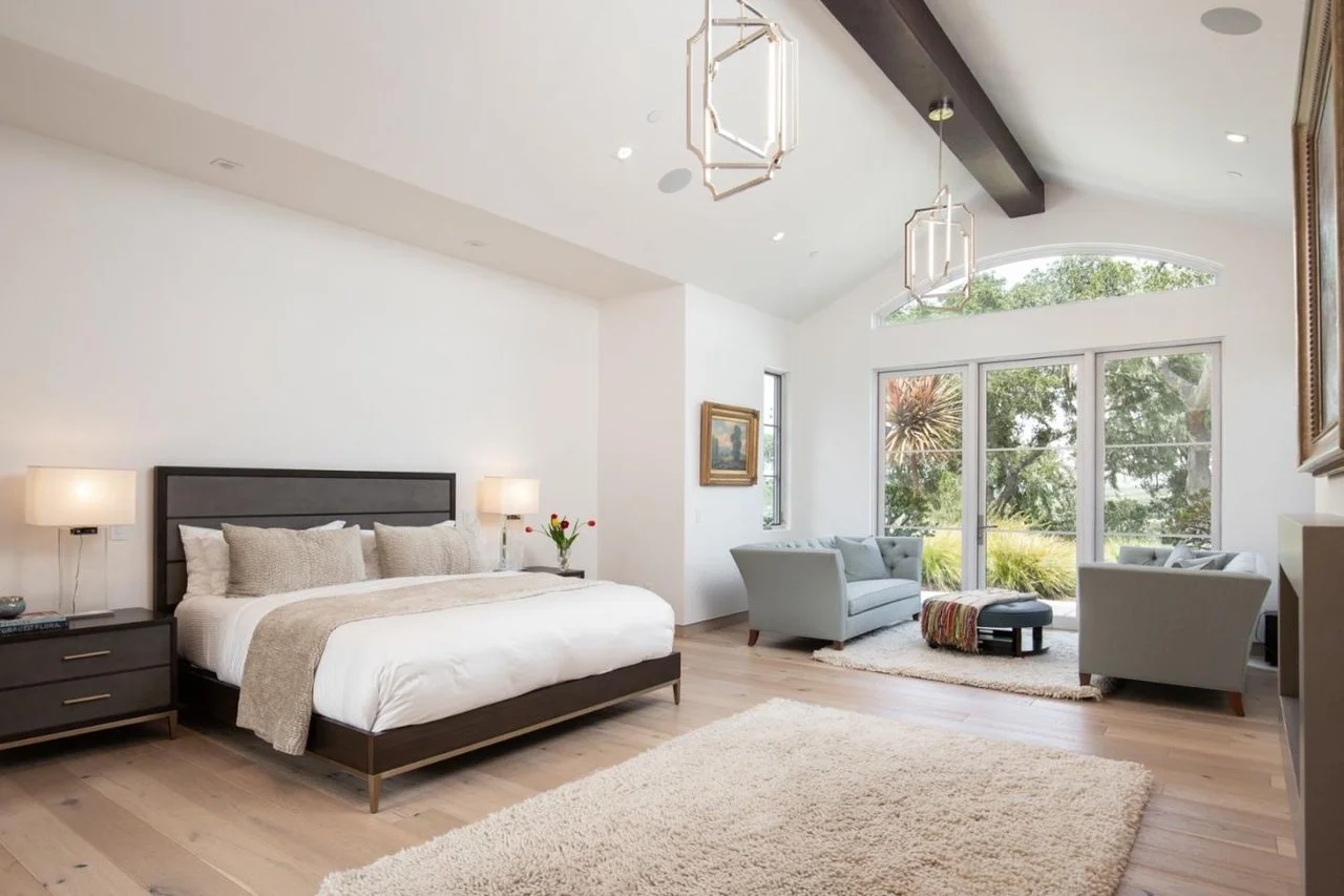 A spacious, modern bedroom with a large bed, two bedside tables with lamps, and a seating area with two armchairs and a window view of trees and greenery.
