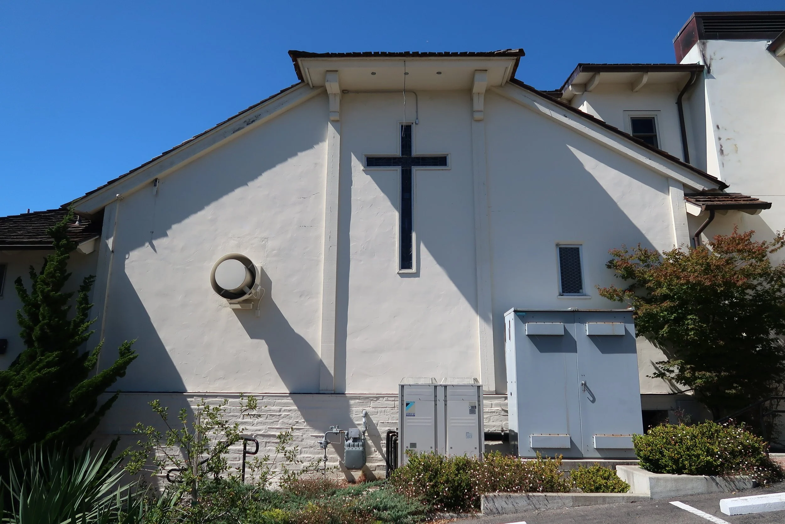 The exterior of a white church building with a cross-shaped window on the front. The building has a sloped roof with brown shingles and is surrounded by small trees and shrubs. There is a utility box and some pipes along the base of the wall.