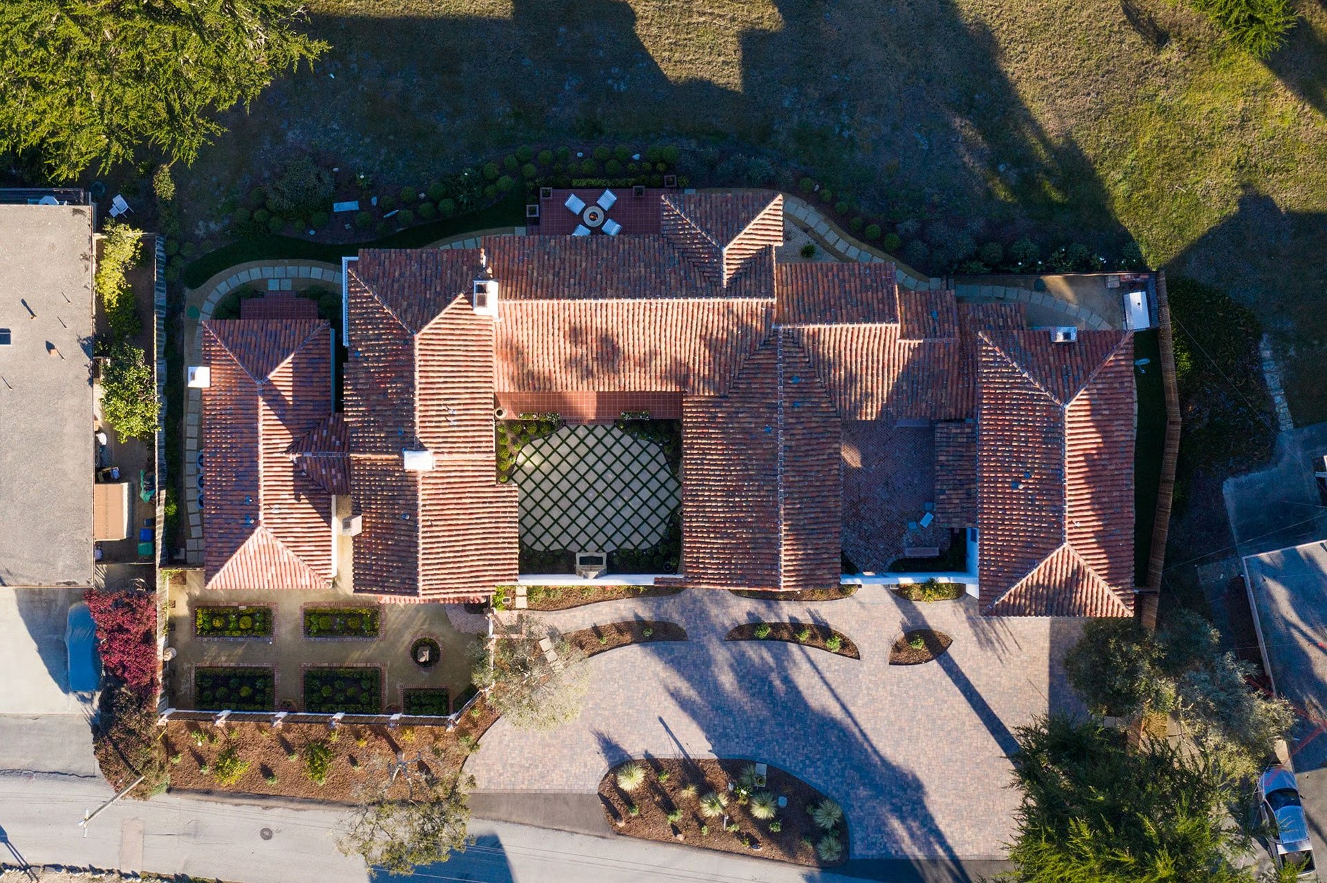 An aerial view of a large house with red-tiled roof, surrounded by landscaped garden and curved pathways, with a street and sidewalk in front and trees casting shadows.