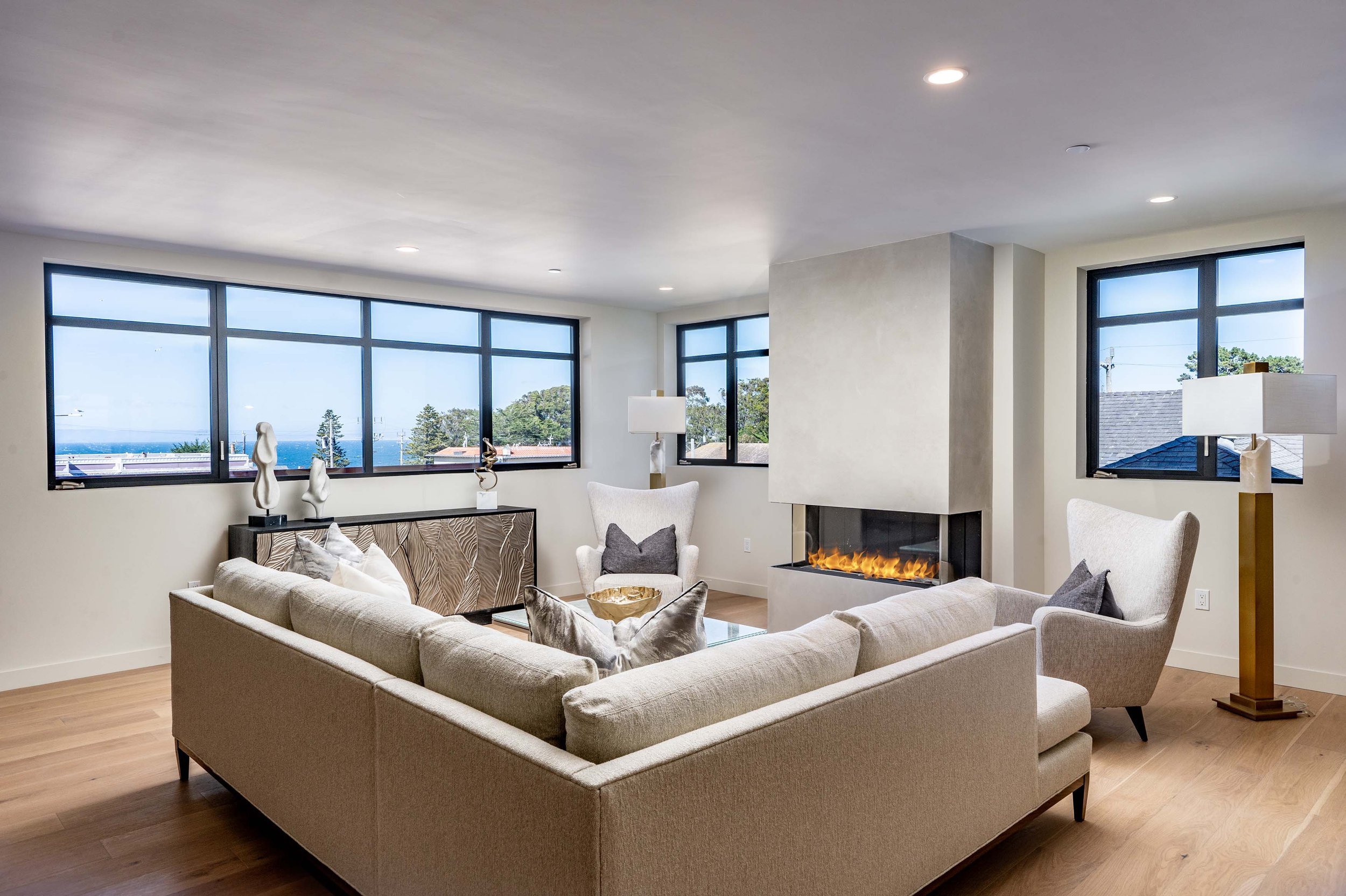 Modern living room with large windows offering ocean and greenery views, a fireplace, and beige furniture.