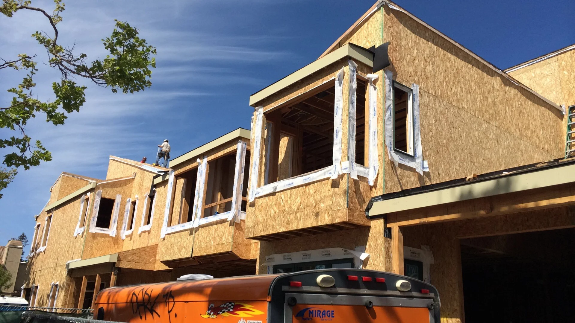 A multi-story residential building under construction with exposed wooden framework and sheathing. Two construction workers wearing hard hats work on the roof. A tree with green leaves is on the left side, and a bright blue sky is in the background.