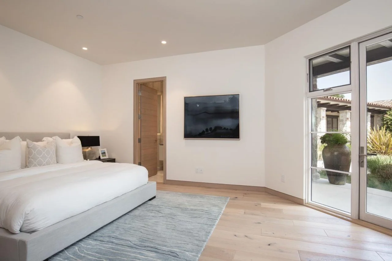 Modern bedroom with white walls, a large bed with white bedding, a black side table with a reading light and framed photo, a wall-mounted TV, a large window with glass door leading outside, and a light-colored wooden floor.