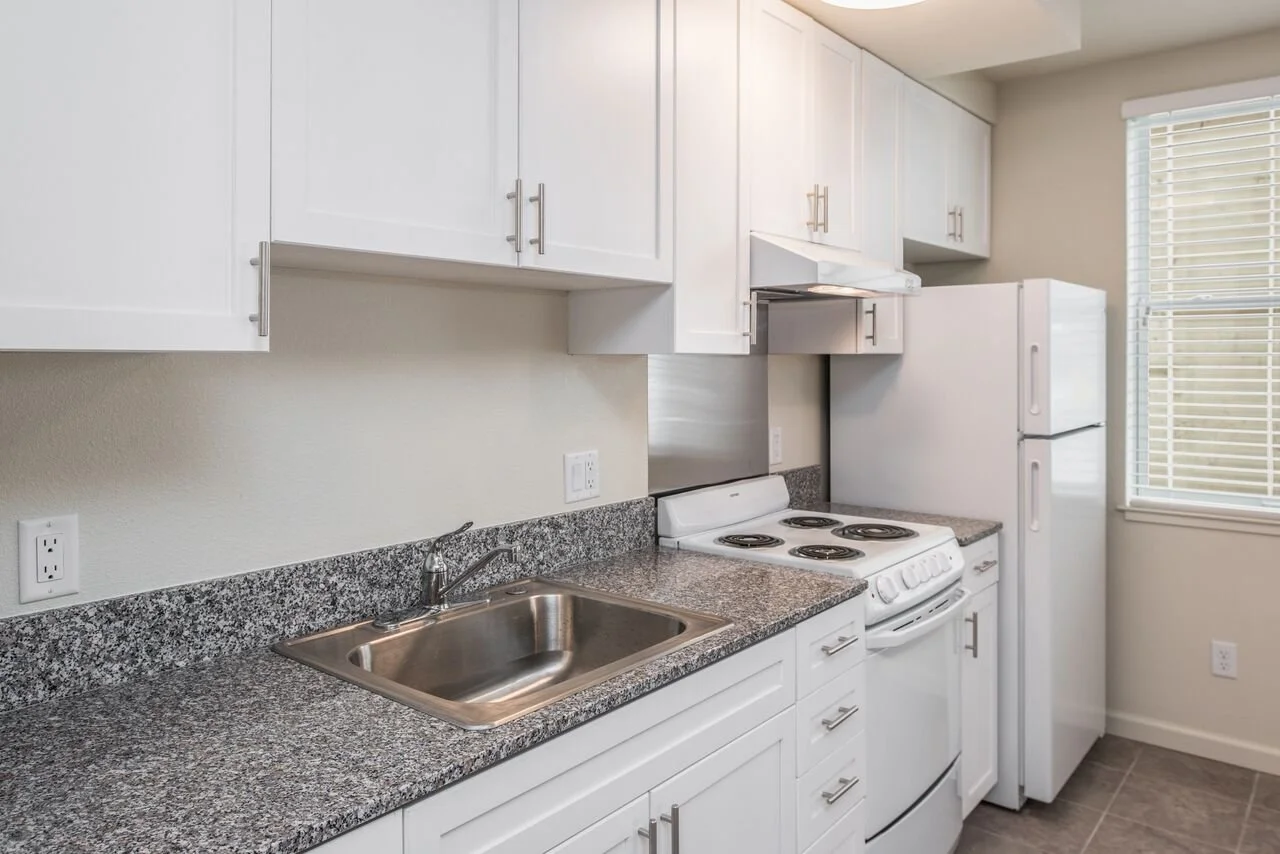 Kitchen with granite countertop, white cabinets, stainless steel sink, white stove, and white refrigerator, near a window with blinds.