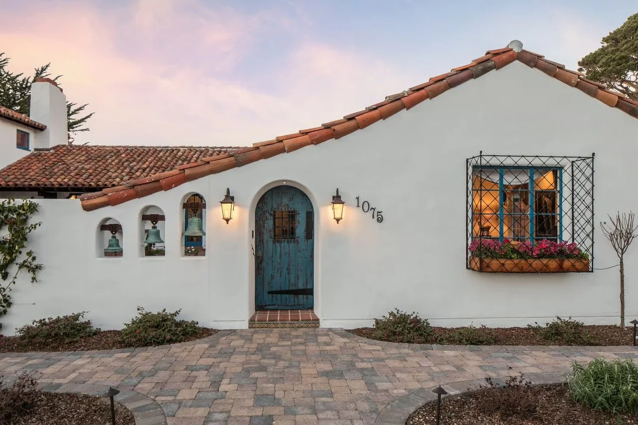 A white stucco house with a red tile roof, blue door, and black wrought iron window grille. The house has two lantern-style lights near the door and a flower box with pink blooms beneath the window. There is a paved walkway and some bushes in front a