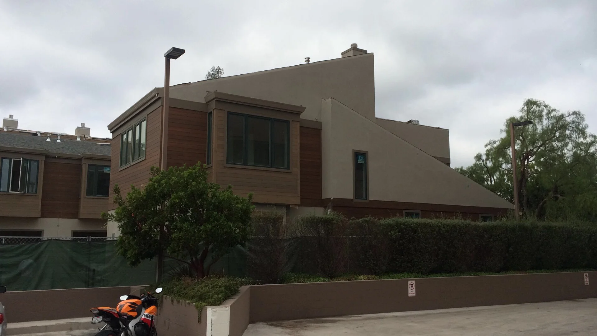 A multi-story residential building with a combination of wood and stucco exterior, large windows, and a sloped roof, situated on a cloudy day with trees and street lamps nearby.