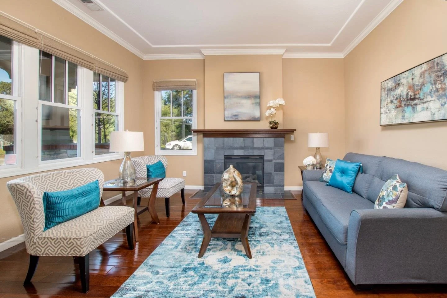 Living room with beige walls, multi-pane windows, a gray brick fireplace, a gray sofa with colorful pillows, a wooden coffee table, two side tables with lamps, artwork on the walls, and a blue patterned rug.
