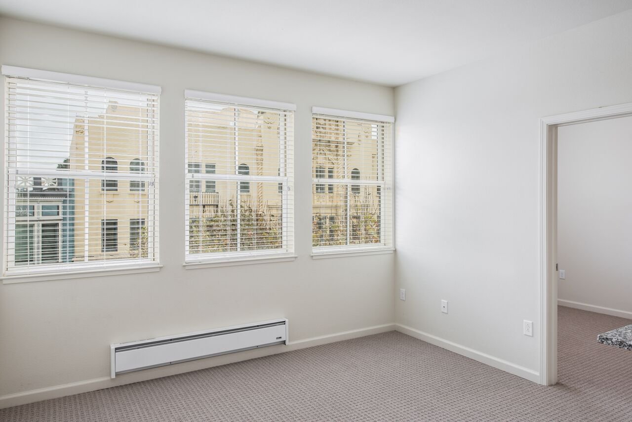 Empty room with three large windows with blinds, white walls, beige carpet, and a baseboard heater.