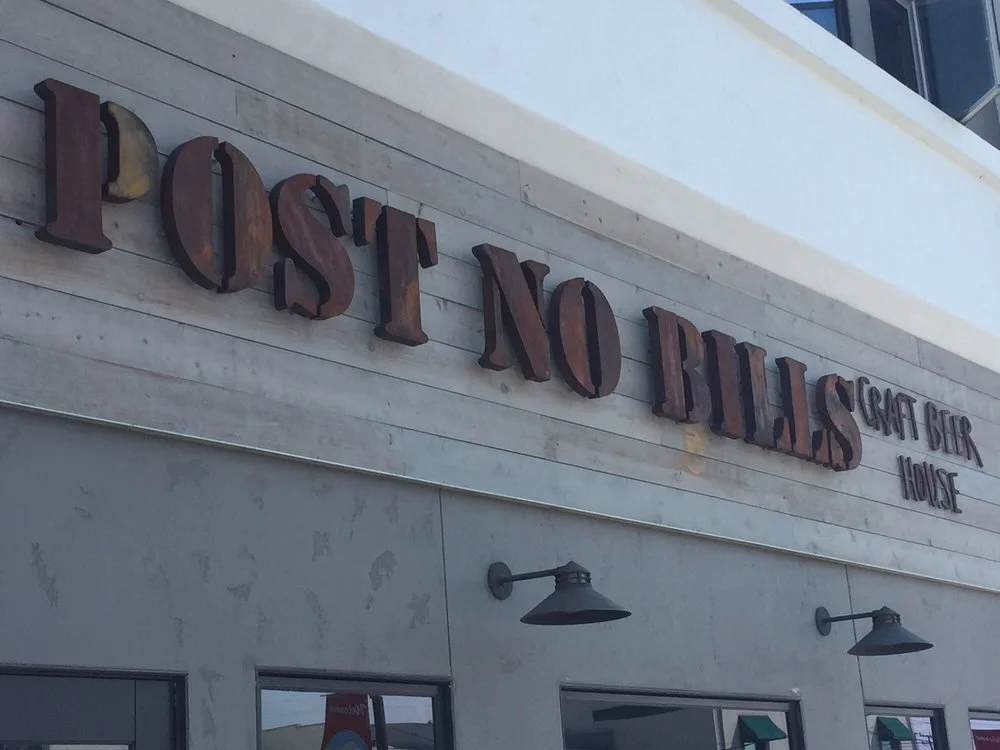 Exterior of a restaurant called 'Post No Bills' with signage indicating craft beer, house, and other offerings, including two black outdoor wall lamps.