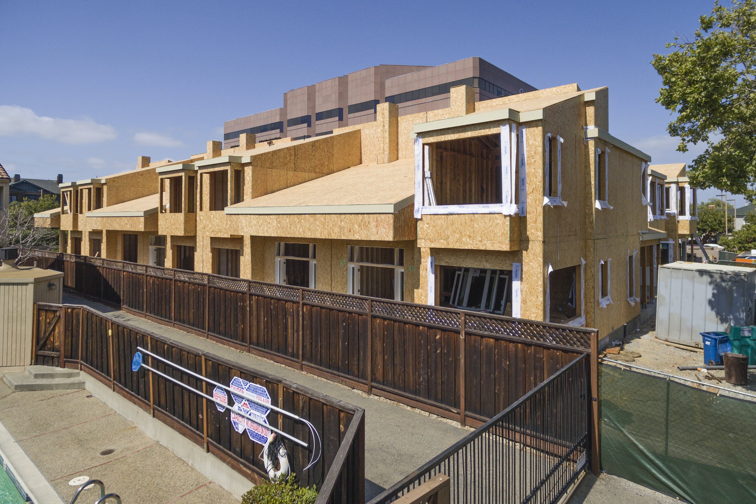 Under construction multi-story residential building with unfinished walls, windows, and exposed wooden framing, surrounded by a safety fence under a blue sky.