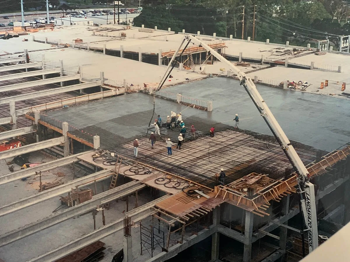 Construction workers pouring concrete on a building's top floor, using a crane to place the concrete.