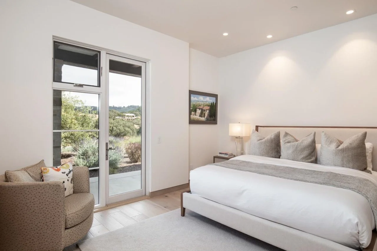 Modern bedroom with a large bed, gray pillows, a side table with a lamp, a framed painting, a beige armchair with a decorative pillow, and a sliding glass door leading outside to a green landscape.