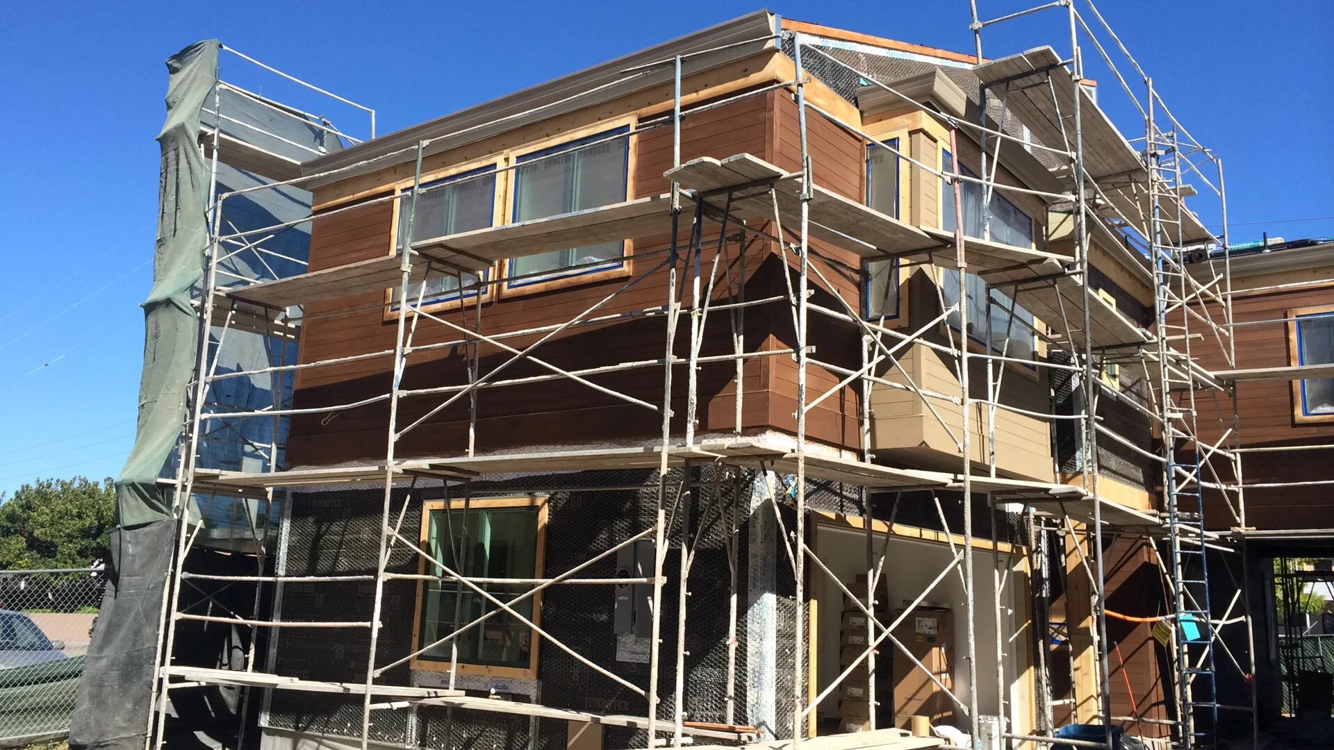 Construction scaffolding surrounds a multi-story building under construction with wooden exterior panels, large windows, and a clear blue sky in the background.