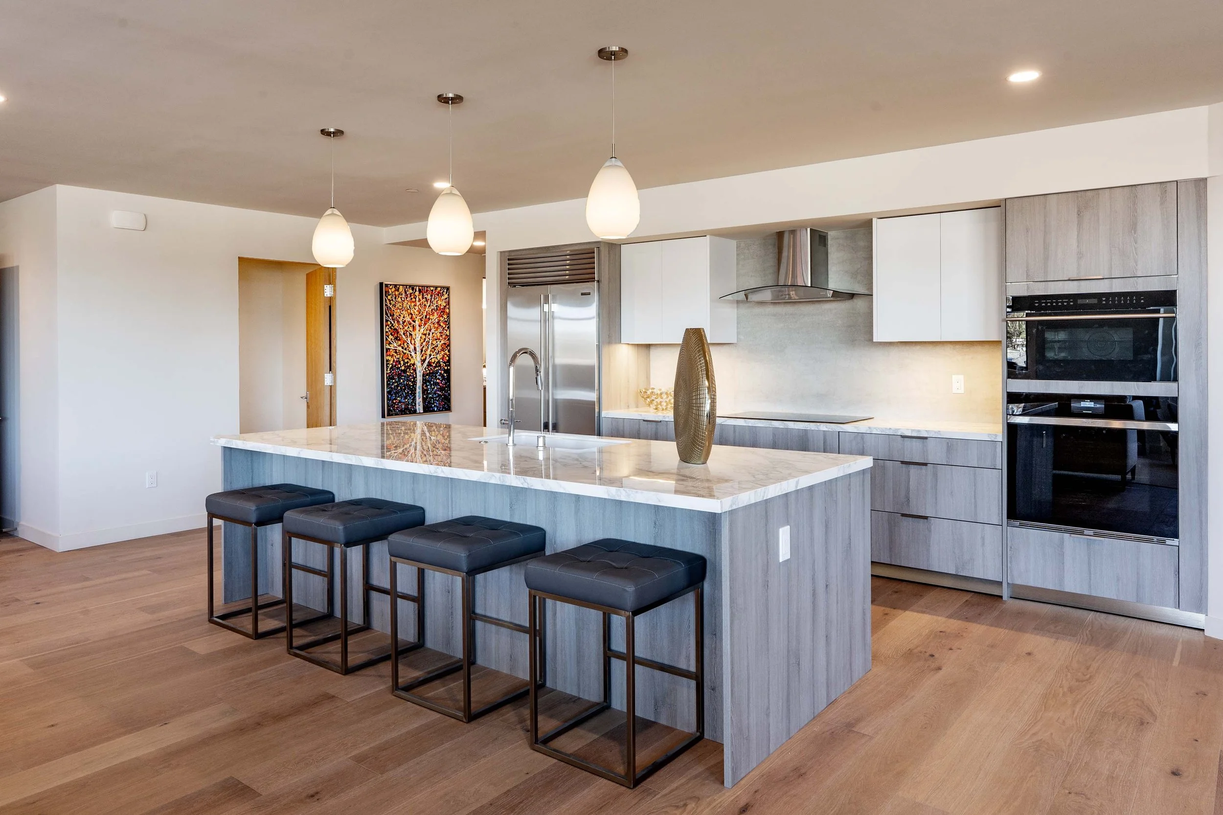 Modern kitchen with white cabinets, stainless steel refrigerator, and a marble island with black stools, opening to a living area with hardwood floors and abstract art.