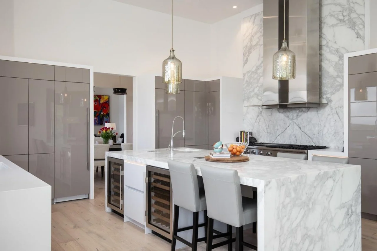 Modern kitchen with white marble island, gray cabinets, stainless steel appliances, pendant lights, and light wood flooring.