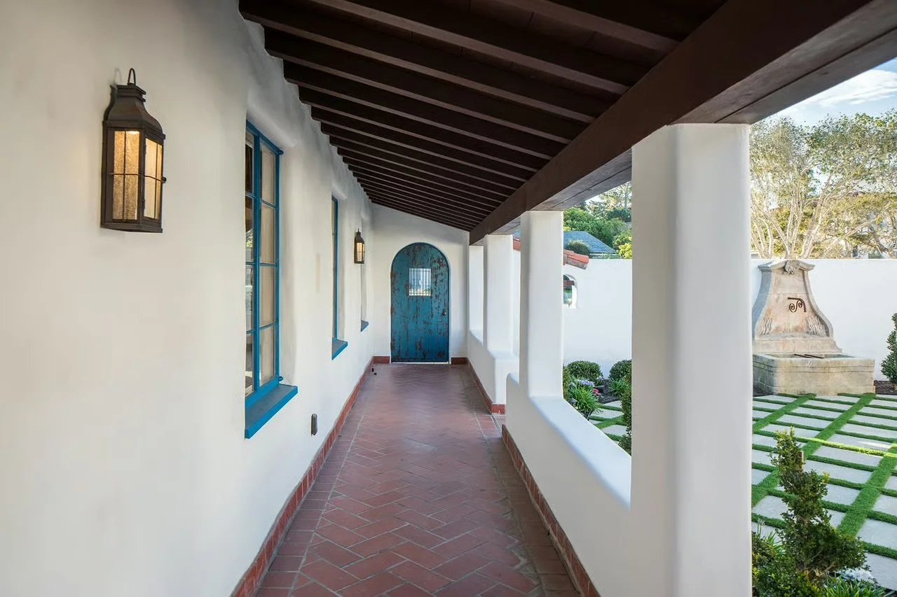 A covered porch with white stucco walls, blue window frames, black lantern-style wall lights, and a rustic blue wooden door with a small window at the end. The porch has a red tile floor, and the area outside features a well-maintained garden with gr