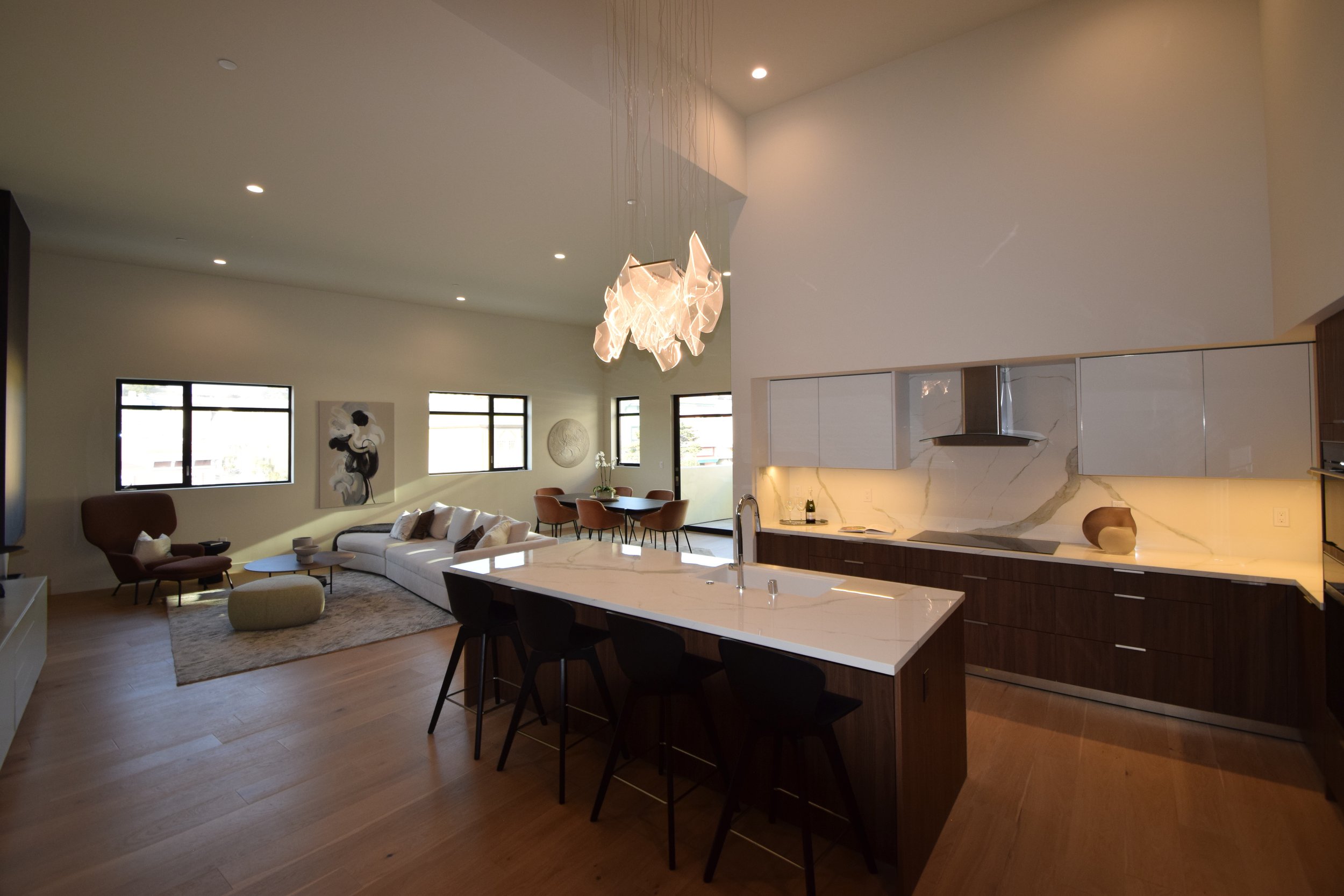 Modern open-concept living space with a kitchen, dining, and living area. Features a white kitchen island with bar stools, a white sofa, armchairs, large windows, and contemporary art on the walls.