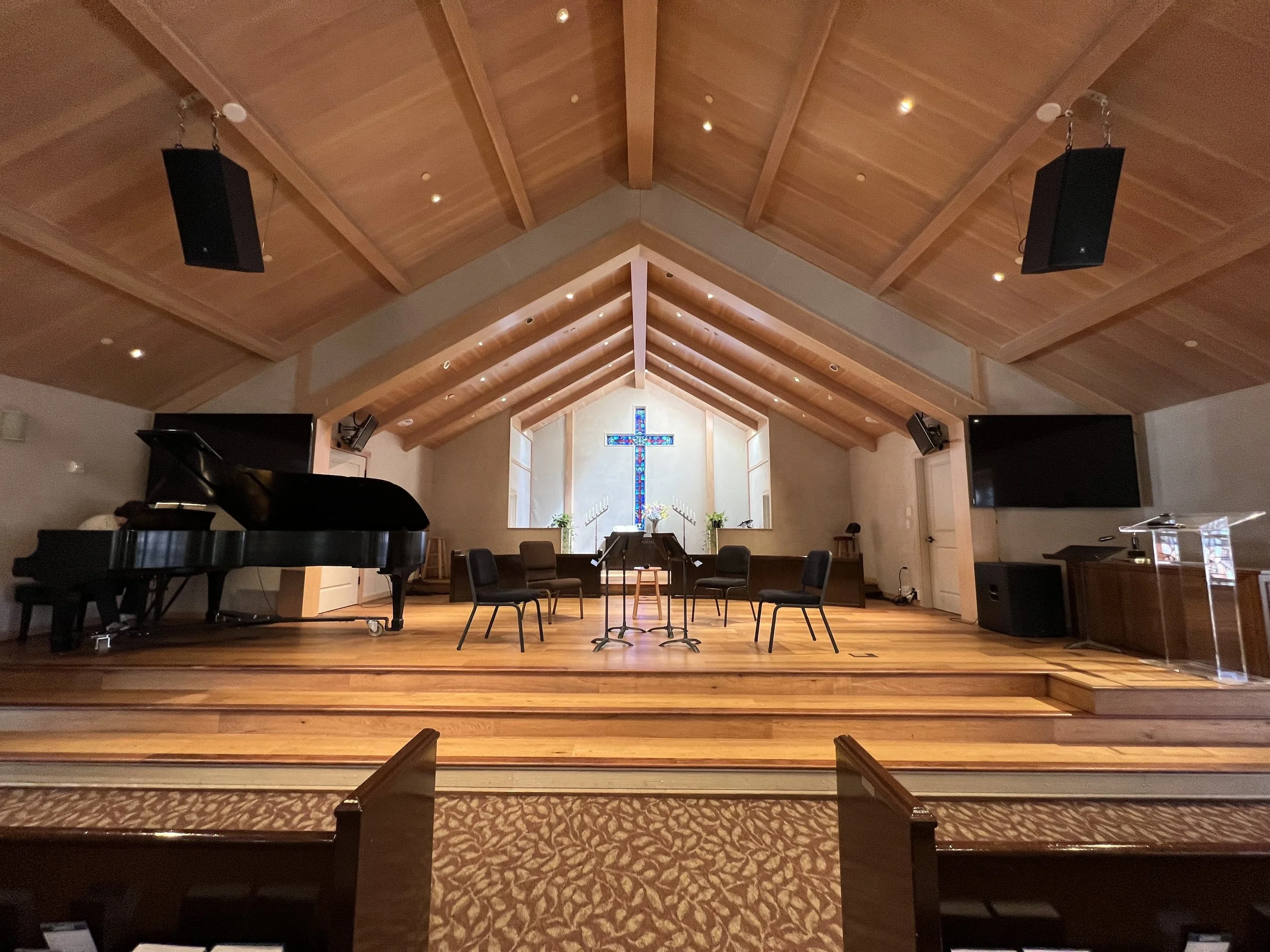 Interior of a church with a wooden ceiling, a stained glass cross at the altar, a piano on the left, and chairs arranged in front.