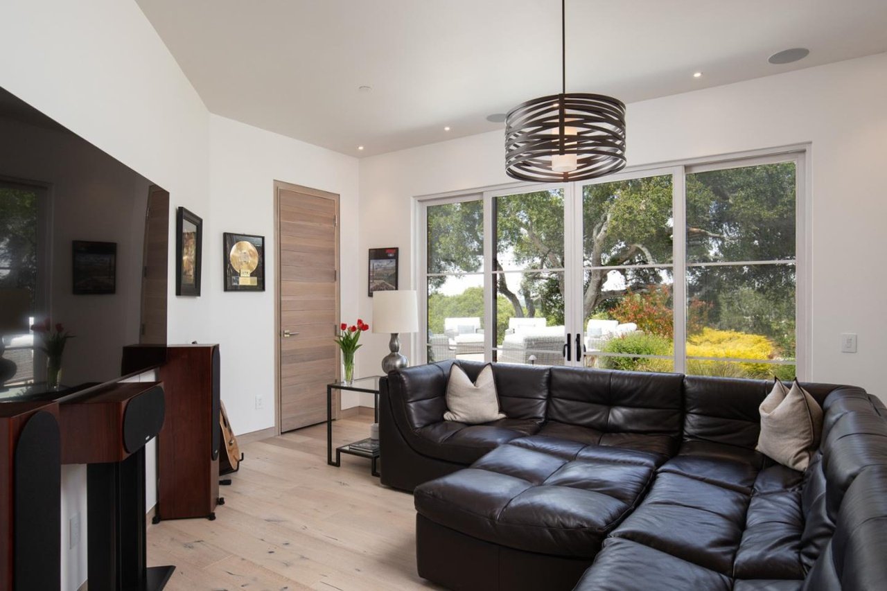 Living room with large sliding glass door, black leather sectional sofa with throw pillows, a side table with a lamp and flowers, wall art, flat-screen TV, and a view of trees and outdoor furniture outside.