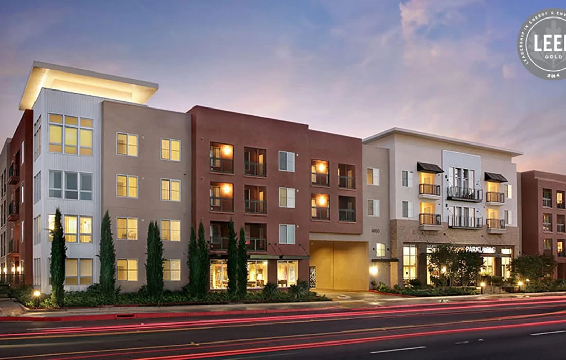 Residential apartment building with multiple stories, windows, balconies, and lit interior lights during dusk, with trees and a streaked light trail from passing cars in the foreground.