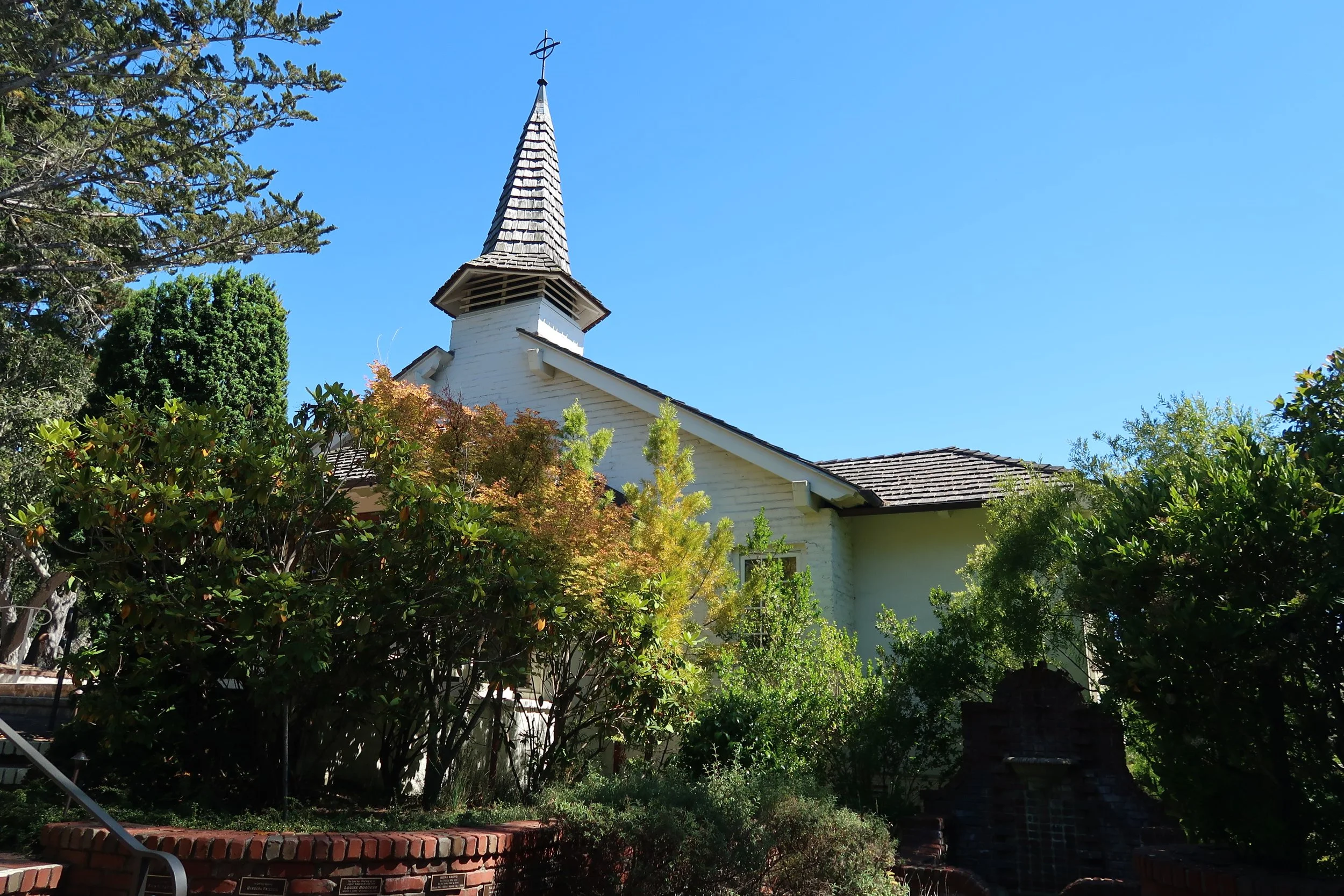 A white church with a tall, narrow steeple topped with a cross, surrounded by green trees and foliage under a blue sky.