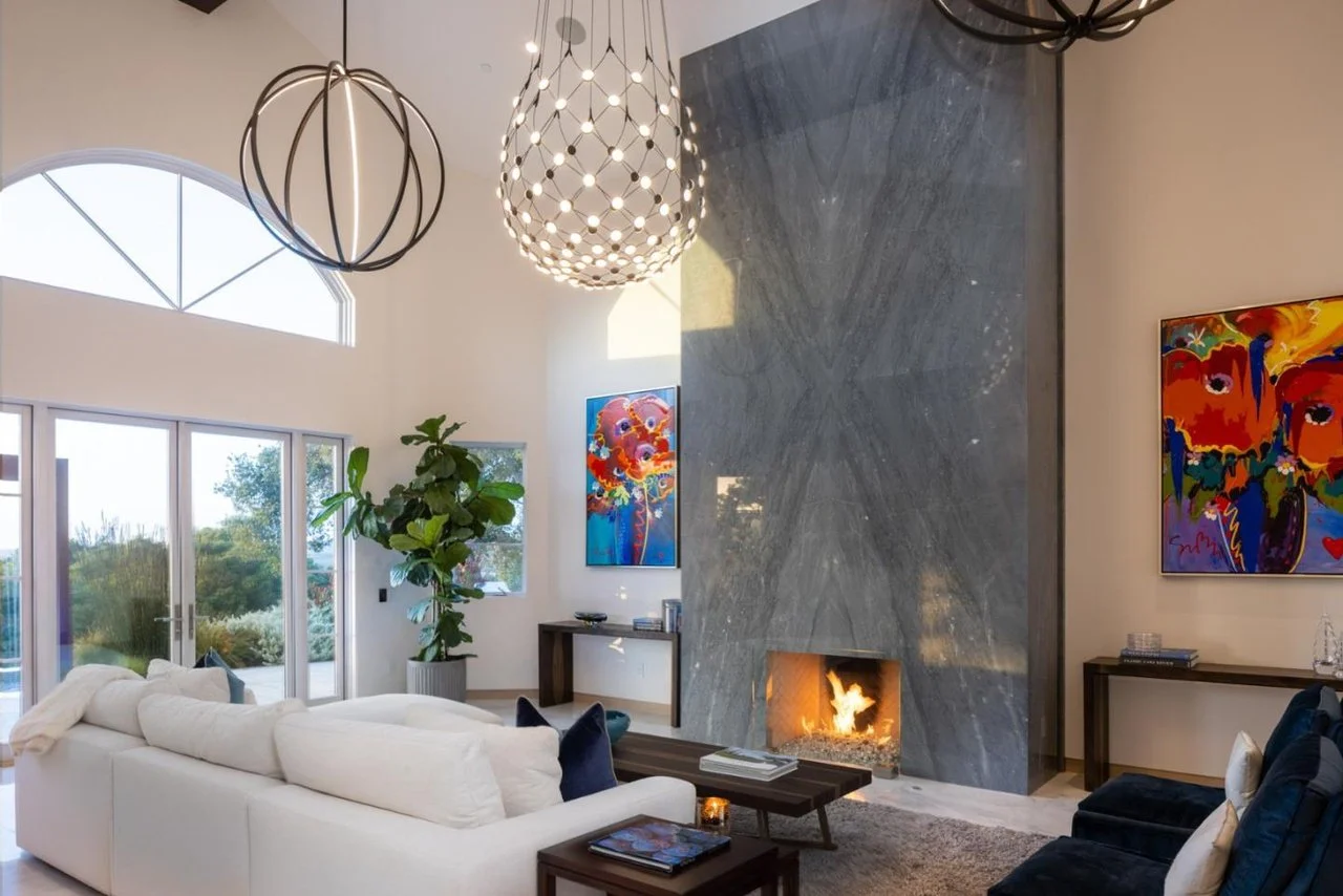 Modern living room with a white sectional sofa, a fireplace, colorful abstract paintings, large windows with outdoor view, and decorative pendant lights.