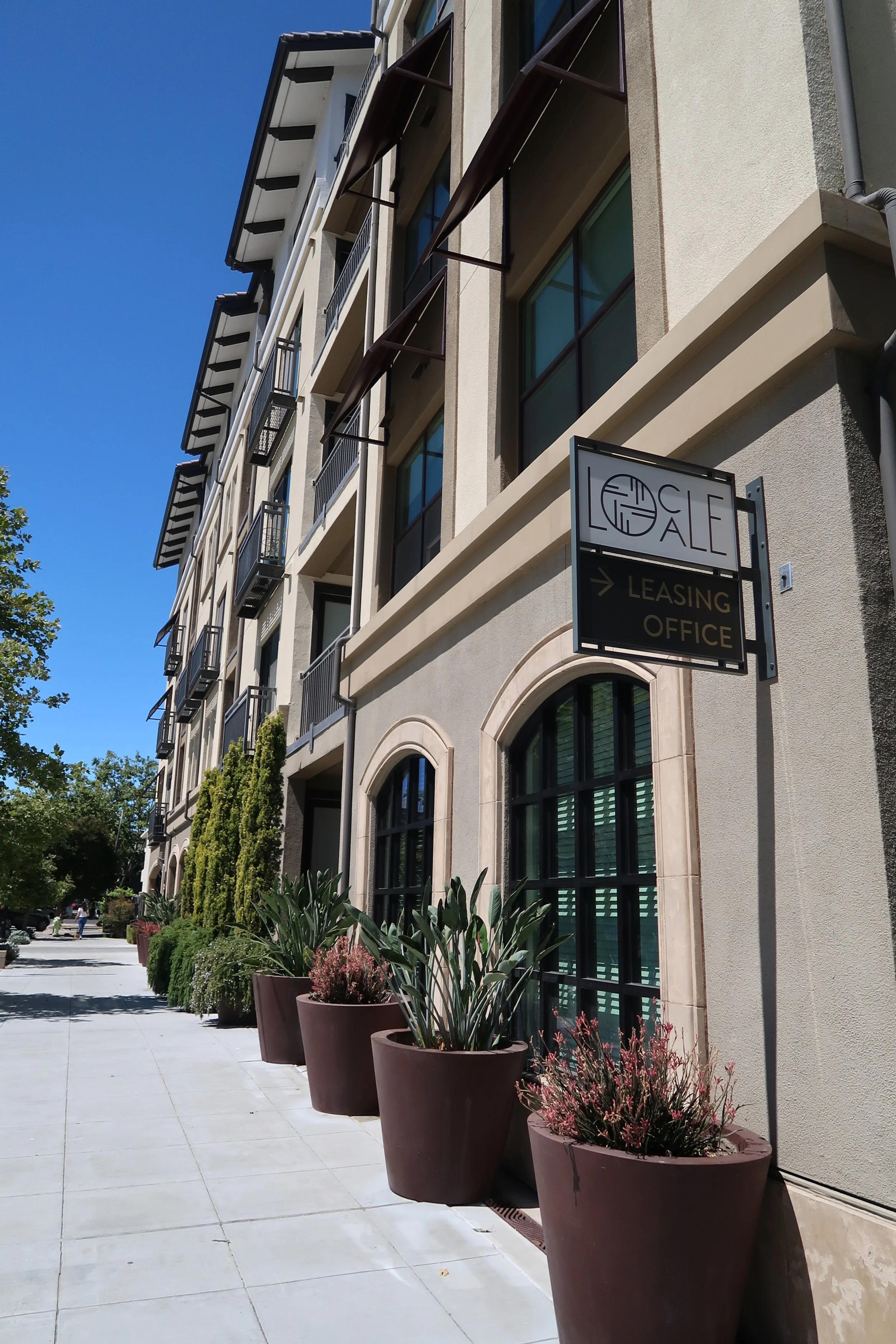 A city street scene with a beige building featuring large windows and black balconies, lined with potted plants on the sidewalk, under a clear blue sky.