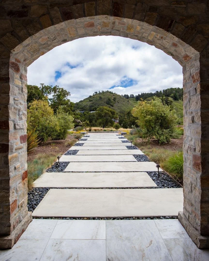 View through a stone archway of a modern pathway with concrete stepping stones and gravel borders, leading to a hilly landscape with trees and a cloudy sky.