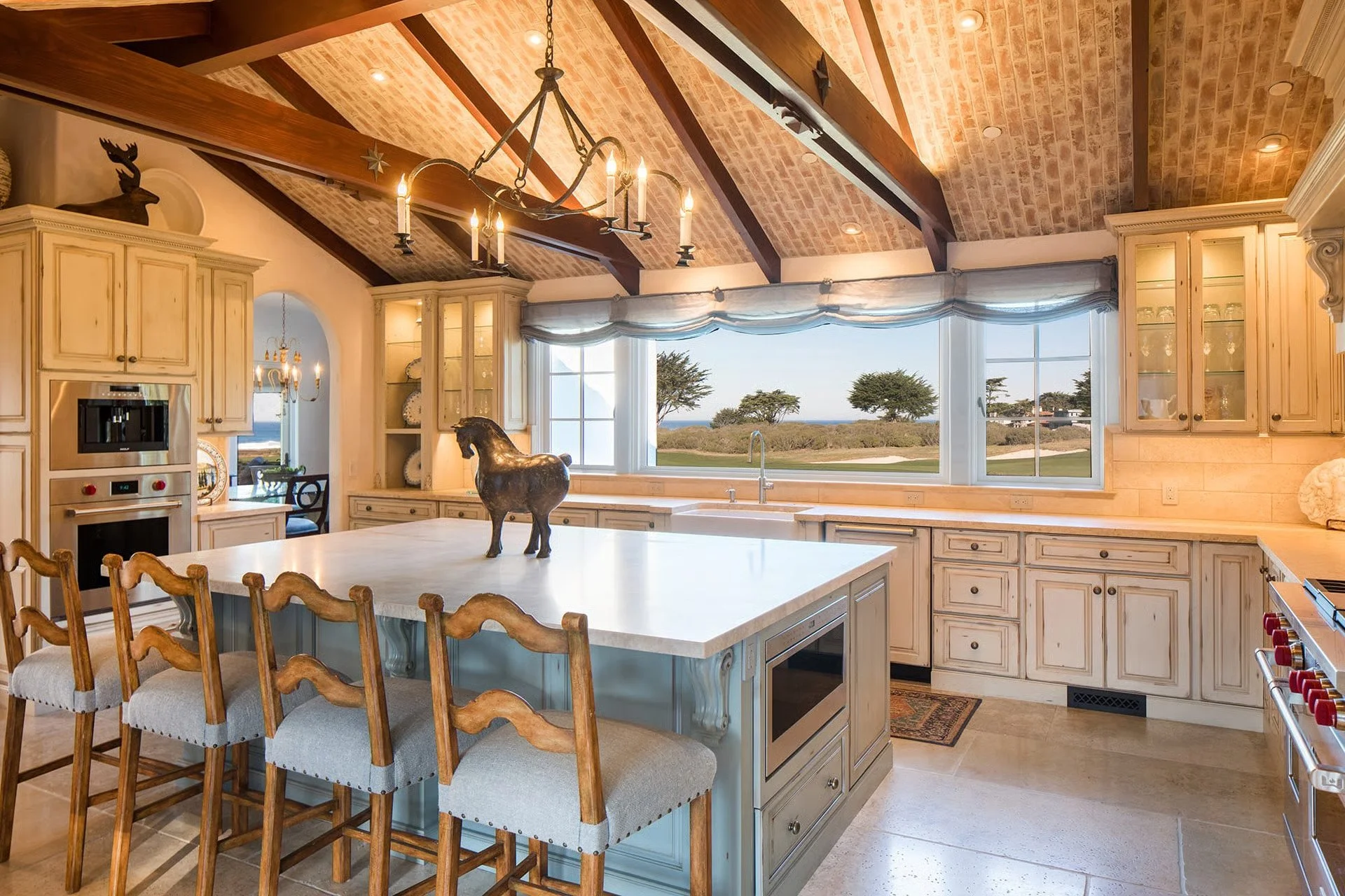 A spacious kitchen with a large island, wooden chairs, white cabinetry, and a view of a landscape through large windows. Decorative items include a small horse sculpture on the island and a moose head decoration on top of a cabinet. The ceiling has e
