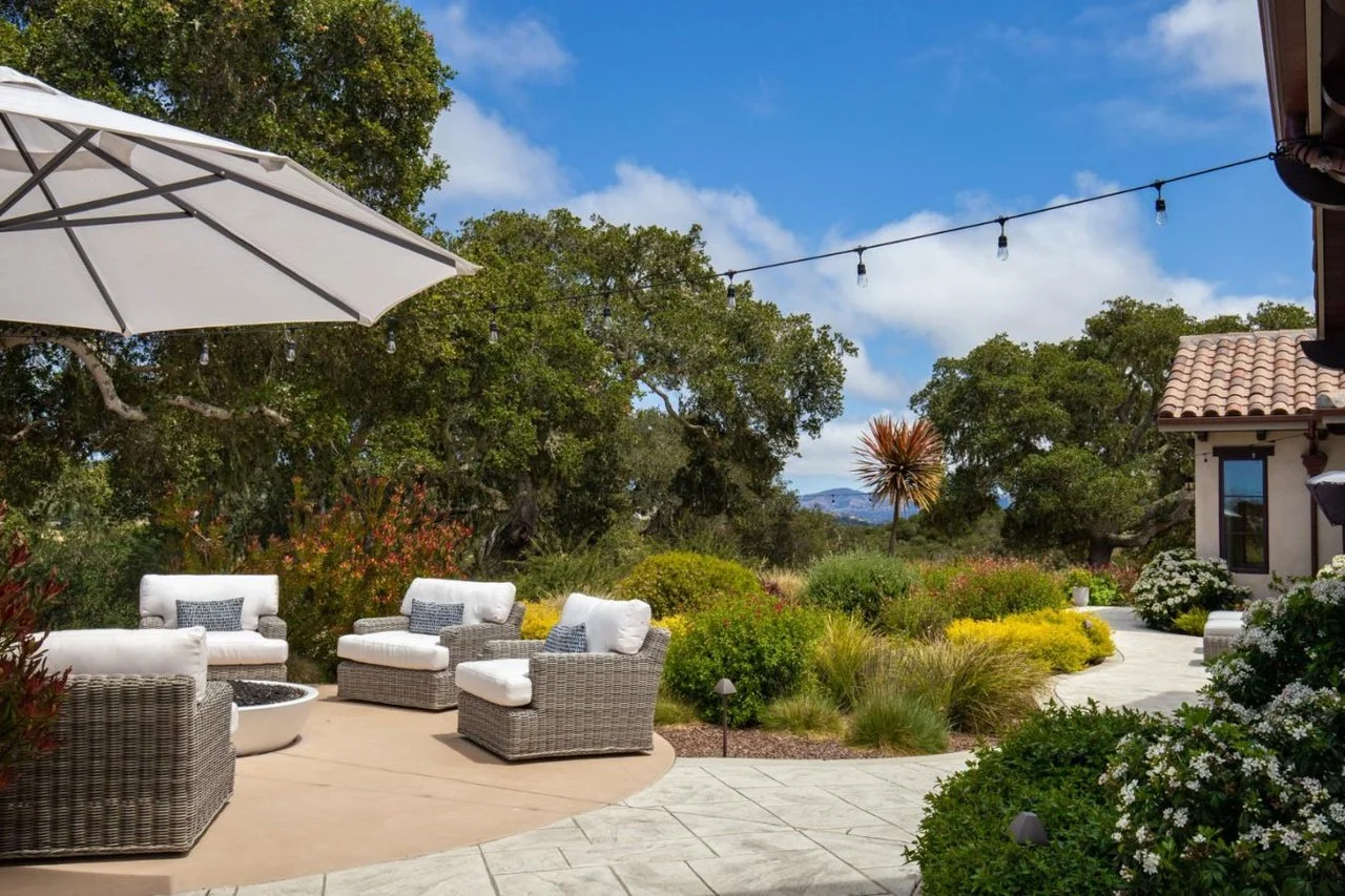 Outdoor patio with wicker furniture and white cushions, surrounded by lush greenery and blooming plants, under string lights and a large umbrella on a sunny day.